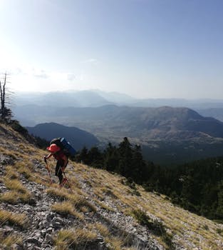 A hiker climbs a rocky mountain trail under bright daylight, with expansive scenic views.